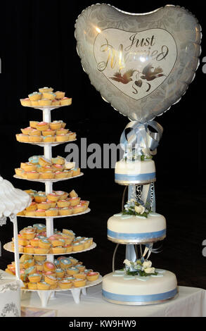 Una torta stand pieno di tortine e una torta di nozze in tre livelli con un palloncino al di sopra con le parole appena sposato scritto su di esso. Catering per matrimoni. Foto Stock