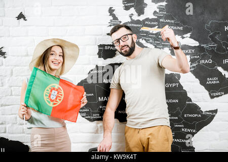 Coppia di giovani viaggiatori in piedi con la bandiera portoghese vicino alla parete con la mappa del mondo, sognando le vacanze estive in Portogallo Foto Stock