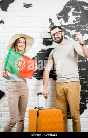 Coppia di giovani viaggiatori in piedi con la bandiera portoghese vicino alla parete con la mappa del mondo, sognando le vacanze estive in Portogallo Foto Stock