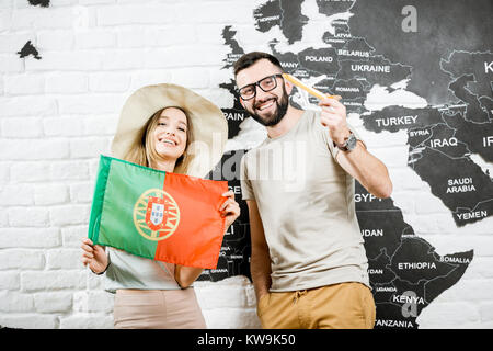 Coppia di giovani viaggiatori in piedi con la bandiera portoghese vicino alla parete con la mappa del mondo, sognando le vacanze estive in Portogallo Foto Stock