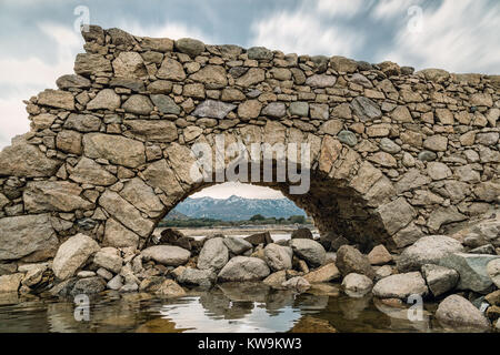 Vista delle montagne coperte di neve attraverso l'arco di un piccolo ponte in pietra a Lac de Codole nella regione della Balagne in Corsica Foto Stock