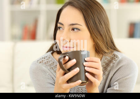 Ritratto di una donna pensieroso tenendo una tazza da caffè guardando il lato seduta su un divano nel soggiorno di casa Foto Stock