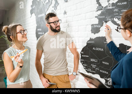 Coppia giovane di viaggiatori scegliendo un viaggio estivo con agente di viaggio puntando sulla mappa del mondo Foto Stock