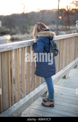 Donna in cappotto invernale cercando sulla ringhiera del piede legno ponte nella foresta al tramonto Foto Stock