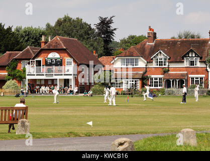 Bowler and fielders in azione in un inglese di cricket con padiglione e case intorno al green Foto Stock