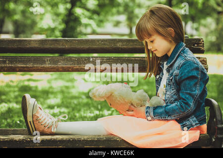 Piccolo felice ragazza seduta su una panchina nel parco e tenendo in mano la sua preferita giocattolo imbottito Foto Stock