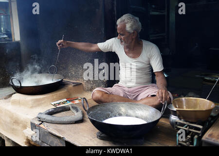 Scena di strada in Pushkar, Senior indian uomo seduto gambe incrociate sul pavimento della sua officina mentre la cottura in pentola grande, Rajasthan, India. Foto Stock