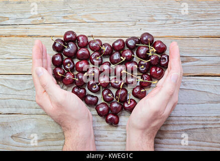 Cuore di fresco e ciliegie mature nel maschio a tazza mani su sfondo di legno Health care nozione di San Valentino e giornata della donna Foto Stock