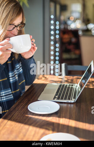 Donna che guarda al computer portatile mentre si sorseggia chai presso la caffetteria in una giornata di sole Foto Stock
