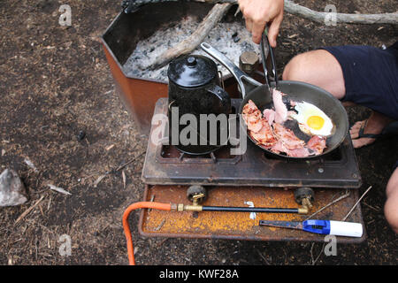 Camping: Uomo cucinare uova e bacon a colazione su un combustibile stufa a campeggio in Australia. Foto Stock