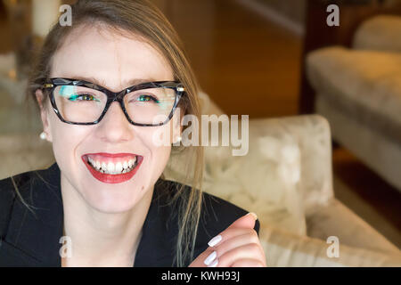 Ritratto di una giovane donna bionda indossando occhiali vista frontale, sorridendo felice, closeup, luce naturale, in interni. Foto Stock