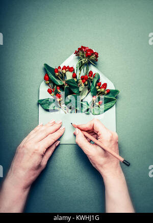 Mani femminili di scrivere con la matita biglietto di auguri su aperto avvolgono rosso con decorazione di autunno bacche e disposizione di fiori su sfondo turchese, parte superiore Foto Stock