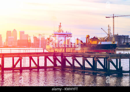 Tramonto la colata di una luce rosa su un molo in disuso adiacente alla Thames Barrier a Londra con un avvicinamento nave e Canary Wharf in background Foto Stock