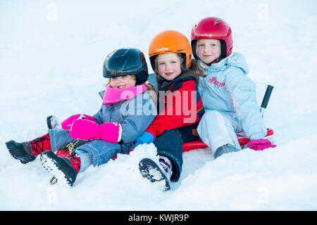 Le ragazze, sorelle, di età compresa tra i tre, cinque e sette anni, e indossano caschi sci per sicurezza, godetevi lo slittino sulla neve fresca su una slitta di colore rosso. (93) Foto Stock