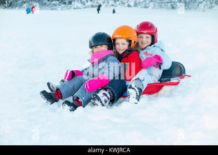 Le ragazze, sorelle, di età compresa tra i tre, cinque e sette anni, e indossano caschi sci per sicurezza, godetevi lo slittino sulla neve fresca su una slitta di colore rosso. (93) Foto Stock