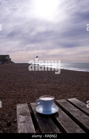 Un bianco tazza da caffè con piattino pieno di caffè cappuccino con cioccolato sulla parte superiore, su di un tavolo di legno su una spiaggia di ciottoli in inverno con il mare, il cielo e Foto Stock