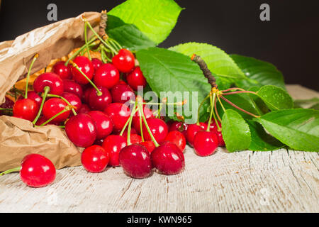 Molti di ciliege rosse con foglie verdi in un sacchetto di carta su un bianco sullo sfondo di legno. Profondità di campo Foto Stock