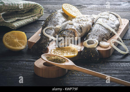 Pesce fresco, posto su di un tagliere di legno e spolverati con erbe aromatiche, fette di limone e cipolla, su un rustico tavolo nero, sotto luce naturale. Foto Stock