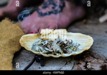 Foto orizzontale di masterizzazione di salvia bianco incenso con flusso di fumo in una conchiglia di mare, sullo sfondo di una donna piede con mandala tattoo, sereno, zen Foto Stock