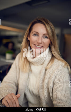 Attraente donna bionda sorridente alla telecamera mentre sorseggiate un bicchiere di vino al chiuso in un pub o ristorante Foto Stock