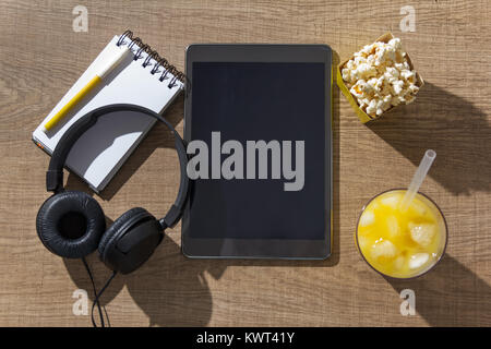 Tavoletta digitale sul tavolo di legno, schermata vuota per il contenuto. Blocco note, auricolari, bicchiere di succo di arancia e pop-corn. Concetto di tempo libero al coperto, il giorno off Foto Stock