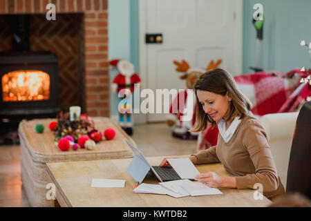 Donna matura è seduta al tavolo da pranzo nella sua casa al tempo di Natale, scrivere cartoline di Natale. Lei sta usando il suo computer portatile per informazioni. Foto Stock