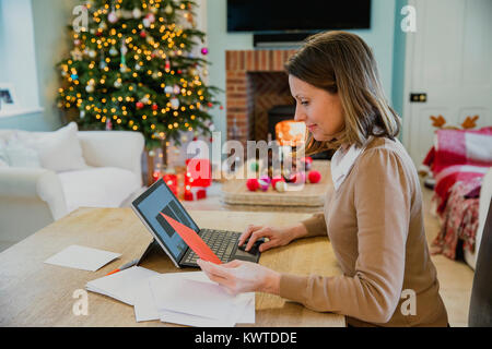 Donna matura è seduta al tavolo da pranzo nella sua casa al tempo di Natale, scrivere cartoline di Natale. Lei sta usando il suo computer portatile per informazioni. Foto Stock