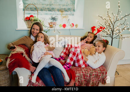 Tre bambine sono rilassanti sul divano con la loro madre al tempo di Natale, guardando la televisione. Foto Stock