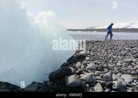 Un grande pezzo di ghiaccio sulla spiaggia Jokursarlon e un uomo a piedi nudi a piedi attraverso acqua fredda della laguna glaciale in Islanda. Foto Stock