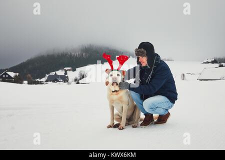 Divertente passeggiata con il cane nel paesaggio innevato. Il Labrador retriever è indossano false corna di renne. La stagione invernale in montagne di Jizera, Repubblica Ceca. Foto Stock
