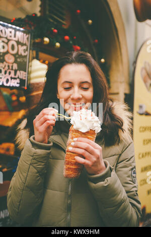 Una bellissima ragazza in una giacca calda mangia trdelnik o Trdlo con crema nelle sue mani, d'inverno in Repubblica Ceca, Praga presso il mercato di Natale. Foto Stock