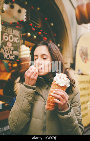 Una bellissima ragazza in una giacca calda mangia trdelnik o Trdlo con crema nelle sue mani, d'inverno in Repubblica Ceca, Praga presso il mercato di Natale. Foto Stock