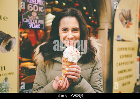 Una bellissima ragazza in una giacca calda mangia trdelnik o Trdlo con crema nelle sue mani, d'inverno in Repubblica Ceca, Praga presso il mercato di Natale. Foto Stock