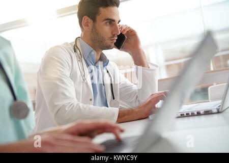 Giovane medico in ufficio a lavorare sul computer portatile e parlando al telefono Foto Stock