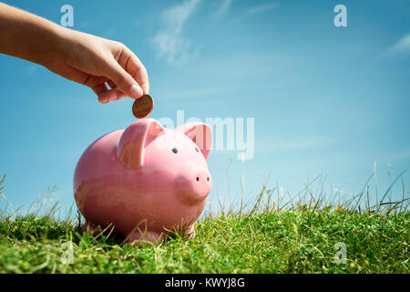 Bambino mano inserire monete e risparmio di denaro nel salvadanaio con erba e cielo blu sullo sfondo Foto Stock