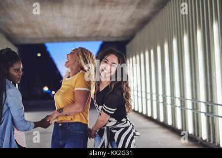Gruppo di diverse giovani donne ridere e camminare mano nella mano insieme verso il basso una passerella nella città di notte Foto Stock
