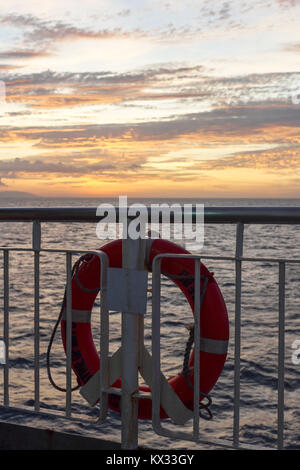 Una luce arancione brillante vita salvagente anello attaccato al corrimano della nave di incrocio tra le isole delle Filippine al tramonto. Foto Stock