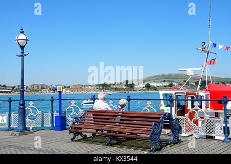 I turisti seduti su una panca in legno lungo il molo vittoriano con vista verso la città, Swanage Dorset, Inghilterra, Regno Unito, Europa occidentale. Foto Stock