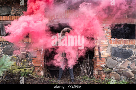 Giovane uomo caucasico in un verde giacca invernale in posa con un rosso bomba di fumo su un triste giorno d'inverno. Tartu, Estonia. Foto Stock