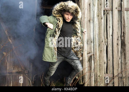 Giovane uomo caucasico in un verde giacca invernale jumping lontano dal fumo di un edificio su un triste giorno d'inverno. Tartu, Estonia. Foto Stock