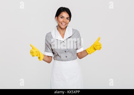 Ritratto di un grazioso gioiosa governante vestito in uniforme che mostra Thumbs up gesto con due mani mentre in piedi e guardando la telecamera isolate su Foto Stock