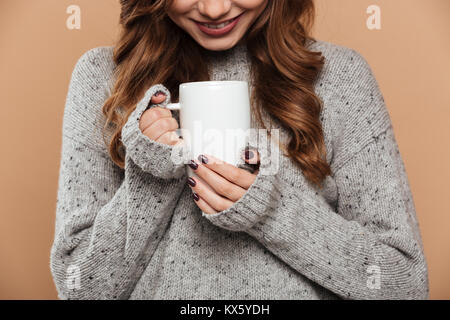 Foto ritagliata di giovani bruna sorridente ragazza in maglia maglione riscaldando le mani tenendo tazza di tè, isolato su sfondo beige Foto Stock