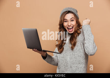 Ritratto di contentissimo brunette girl in maglione di lana pugno di aggraffaggio nel gesto vincitore tenendo laptop, isolato su sfondo beige Foto Stock