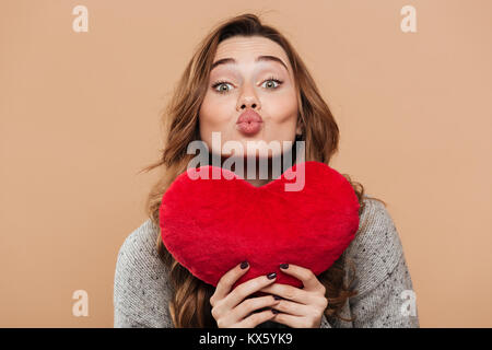 Close-up di foto bella bruna ragazza con grande rosso morbido cuore mentre l'invio kiss, guardando la telecamera, isolato su sfondo beige Foto Stock