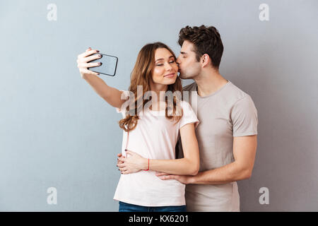 Ritratto di una felice coppia giovane abbracciando e baciando tenendo un selfie oltre il muro grigio Foto Stock