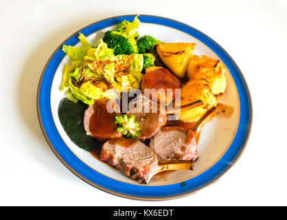 Cena inglese carré d'agnello arrosto Cavolo broccoli patate arrosto salsa alla menta e sugo di carne servita su un blu e piastra bianca su sfondo bianco Foto Stock