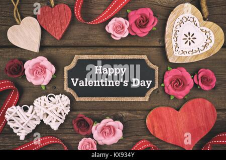 Happy Valentines Day messaggio su un tag di lavagna con cornice di cuori e fiori su un sfondo di legno. Lo stile vintage. Foto Stock