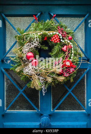 Vista frontale di una corona di Natale tradizionale fatta di rami di pino, palline di Natale rosso lucido e bacche rosse appesi su una porta della casa mezzo-vetrata. Foto Stock