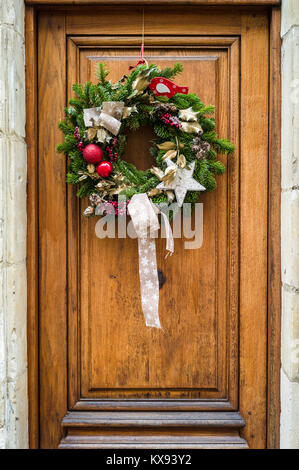 Vista frontale di un tradizionale Natale corona fatta di pino rami, rosso palle di Natale e Holly foglie e bacche appeso su una tavola di legno porta anteriore. Foto Stock