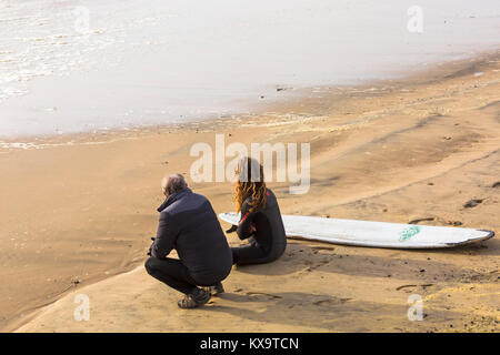 Surfer seduto sulla spiaggia accanto alla tavola da surf con un altro uomo che guarda al mare a Bournemouth Beach, DORSET REGNO UNITO NEL GENNAIO Foto Stock